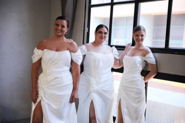 Three brides in satin off-the-shoulder wedding gowns with thigh-high slits, celebrating inclusive sizing and elegant bridal fashion