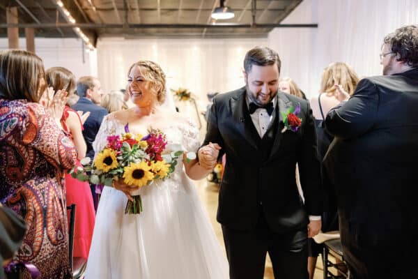 The newlyweds beam with joy as they walk down the aisle, hand in hand, surrounded by cheers and applause. The bride's vibrant sunflower bouquet perfectly complements her radiant smile, while the groom looks dapper in his classic black tuxedo.
