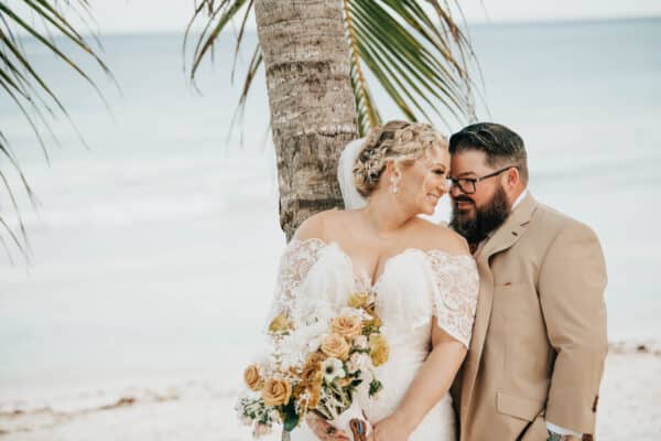 A romantic moment by the beach featuring a bride in a stunning plus-size boho wedding dress with off-the-shoulder lace detailing, holding a bouquet of earthy-toned flowers. Her intricate braided hairstyle complements the serene ocean backdrop as she shares an intimate glance with her groom in a tan suit.