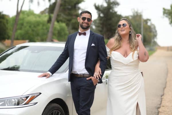 A joyful bride in a stunning off-the-shoulder plus-size wedding dress with a thigh-high slit walks arm-in-arm with her groom, both wearing stylish sunglasses. The sleek white car in the background and the natural surroundings add a modern and relaxed vibe to this memorable moment