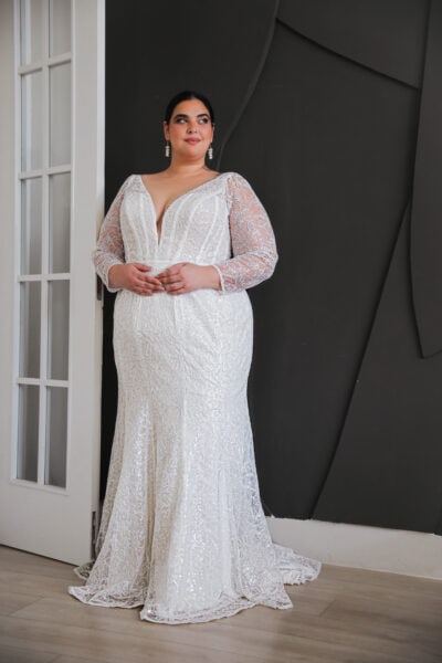 "Plus-size bride wearing a shimmering lace wedding gown with a plunging neckline and long sheer sleeves, standing confidently against a sleek, modern backdrop."