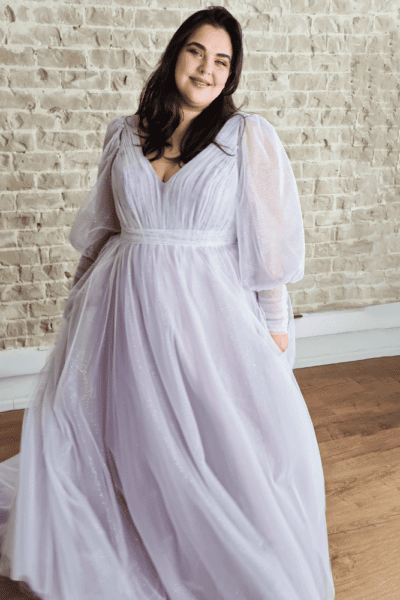 plus-size wedding dress in a soft lavender hue featuring a flowing A-line silhouette with a deep V-neckline. The gown is enhanced by sparkling fabric and elegant puffed long sleeves
