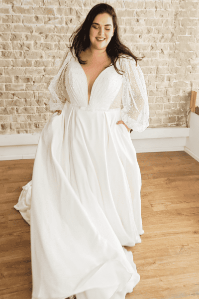 plus-size wedding dress featuring a deep V-neckline and a flowing A-line silhouette. The gown is adorned with intricate beading on the bodice and is complemented by sheer, puffed long sleeves,