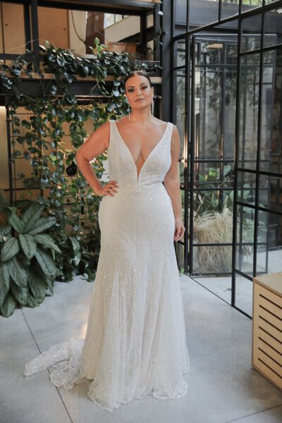 Plus-size model in a beaded, sleeveless wedding dress with a deep V-neckline, posing in a modern, greenery-filled setting.