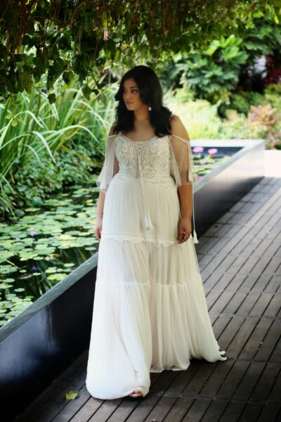 a curvy bride in a flowy boho white dress with off shoulder sleeves standing on a deck