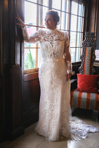 The plus-size bride, wearing a lace gown with a high neckline, long sheer sleeves, and floral embroidery