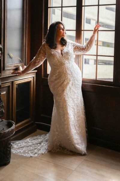 The plus-size bride, adorned in a lace gown with detailed embroidery, long sheer sleeves, and a trailing train