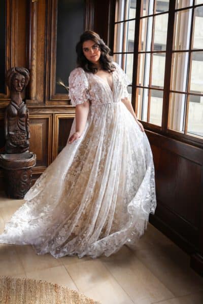 This plus-size wedding gown features voluminous flutter sleeves and an A-line silhouette crafted from floral-embroidered lace. It includes a deep V-neckline and delicate shimmer throughout the fabric, offering an elegant and romantic design suitable for various wedding themes.
