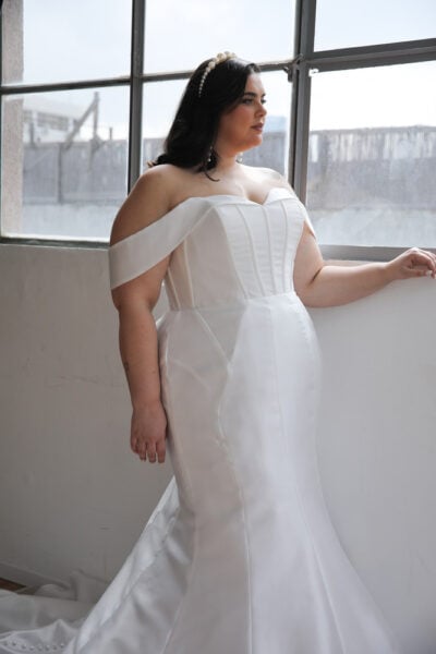 Plus-size bride wearing a stunning plus-size wedding dress featuring an invisible internal corset. The gown has a sleek mermaid silhouette with a fitted bodice, a sweetheart neckline, and elegant off-the-shoulder sleeves.
