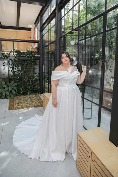 Plus-size bride in a luxurious off-the-shoulder gown with a flowing train, standing in a bright, modern space with glass walls and lush greenery.