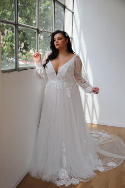 Plus-size bride wearing an enchanting plus-size wedding dress featuring an invisible internal corset. The gown showcases a deep V-neckline with delicate lace detailing and long sheer sleeves. Flowing A-line silhouette