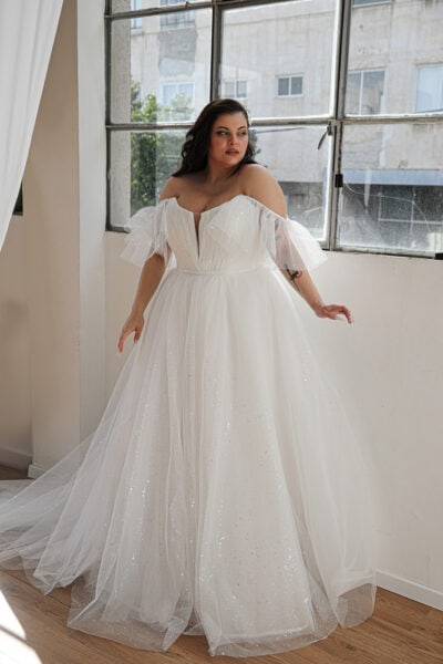 Plus-size bride wearing a stunning plus-size wedding dress with an invisible internal corset. The gown features a sweetheart neckline and flowing A-line silhouette adorned with sparkling tulle. Off-the-shoulder flutter sleeves add a romantic touch
