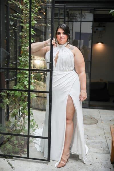 Plus-size bride in a modern halter-neck gown with a thigh-high slit, standing confidently in a glass doorway surrounded by lush greenery.