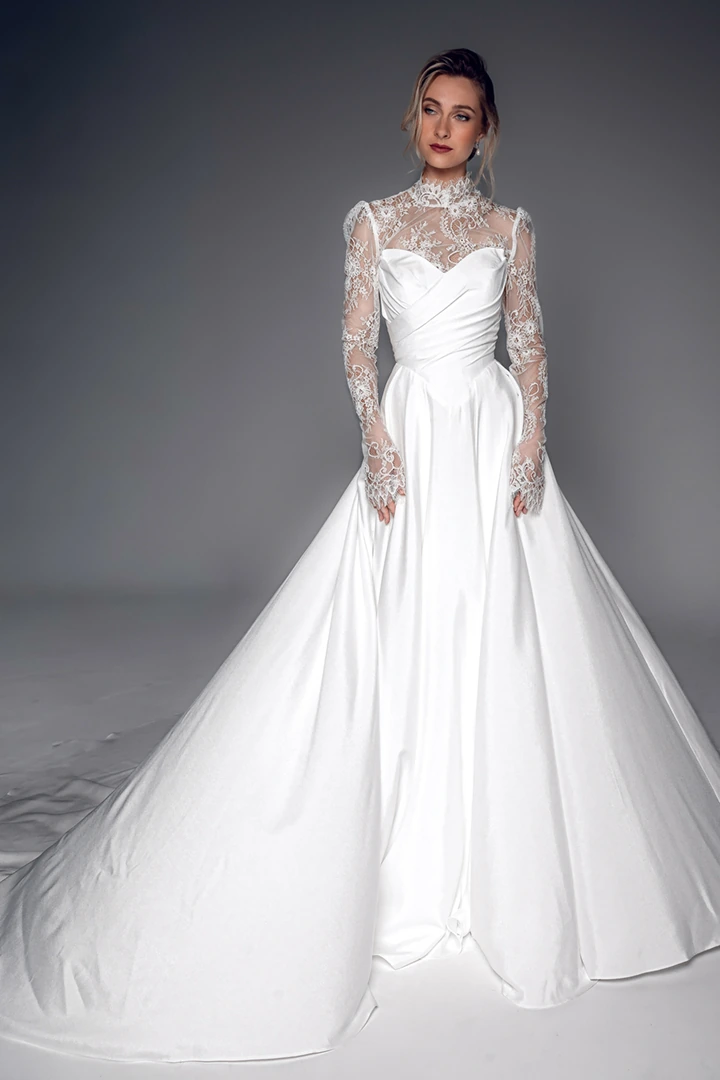 שמלת כלה מנון של סטודיו לבנה בגזרת A, עם חצאית חלקה ומבריקה, מחוך עם קפלים, מחשוף תחרה גבוה ושרוולי תחרה ארוכים, מבט מלפנים באורך מלא.