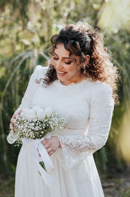 כלה לובשת שמלת כלה צנועה עם שרוולים ארוכים ותחרה עדינה, מחזיקה זר לבן ומביטה בו בחיוך, מצולמת בטבע באור רך