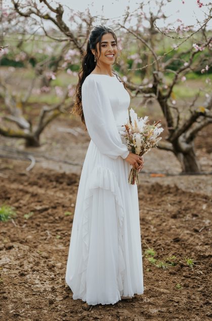 כלה לובשת שמלת כלה צנועה עם שרוולים ארוכים ומחזיקה זר פרחים טבעי, מצולמת בפרדס פורח באווירה כפרית ורומנטית.